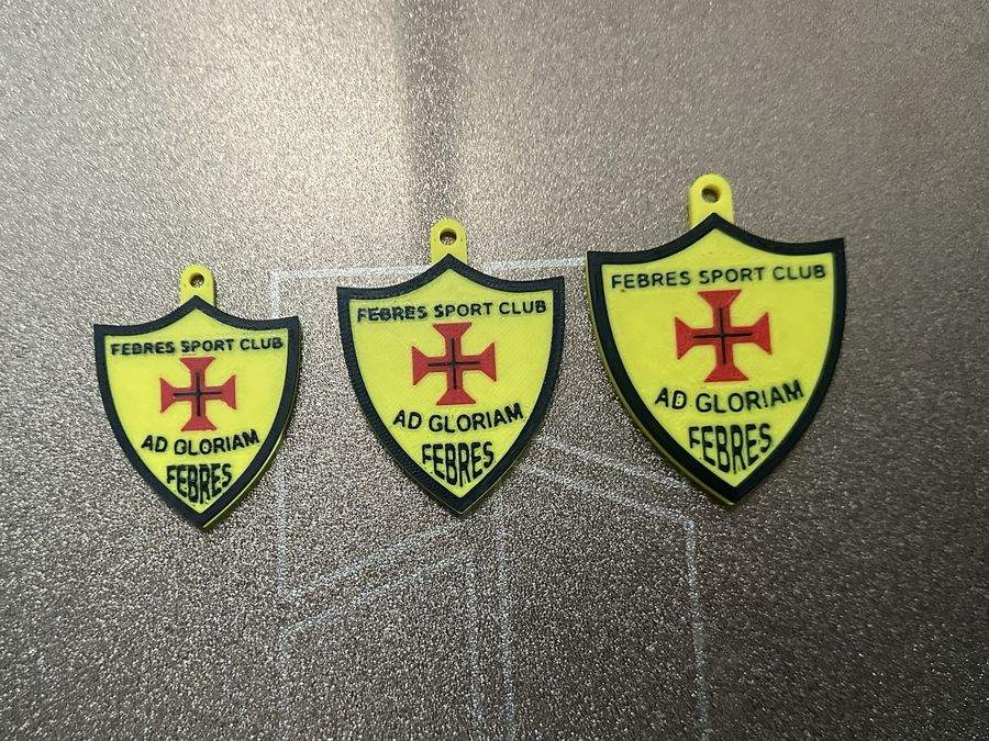 Imagem ilustrativa: três porta-chaves impressos em 3D com um emblema em amarelo, preto e vermelho, de três tamanhos.